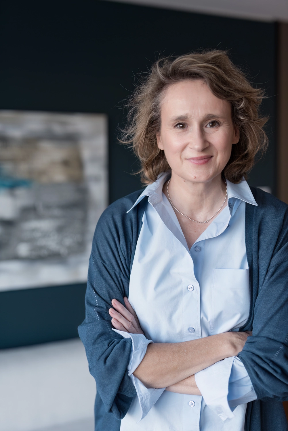 Portrait1  frontal Gesicht und Oberkörper Christiane Schicker vor einer Wand in dunkelblauem Anstrich und der Andeutung eines Wandbildes. CS trägt einen blauen Cardigan und eine hellblaue Hemdbluse