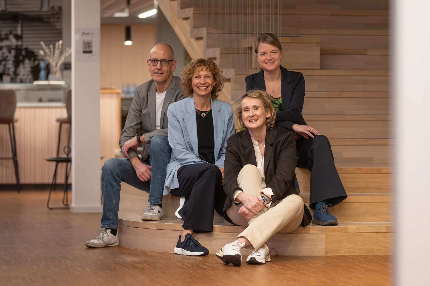 4 Personen sitzen in einem Co-Working-Büroloft, locker gruppiert auf Treppenstufen in einem warmen, hellen Holzton, dem Fotografen zugewandt: von links nach rechts: Gernot Bracher, Inhaber Con4Rent Kirchheim unter Teck, Anja Lippert-Hertle, Christiane Schicker, Saskia Klinger, Wirtschaftsförderung Kirchheim unter Teck
