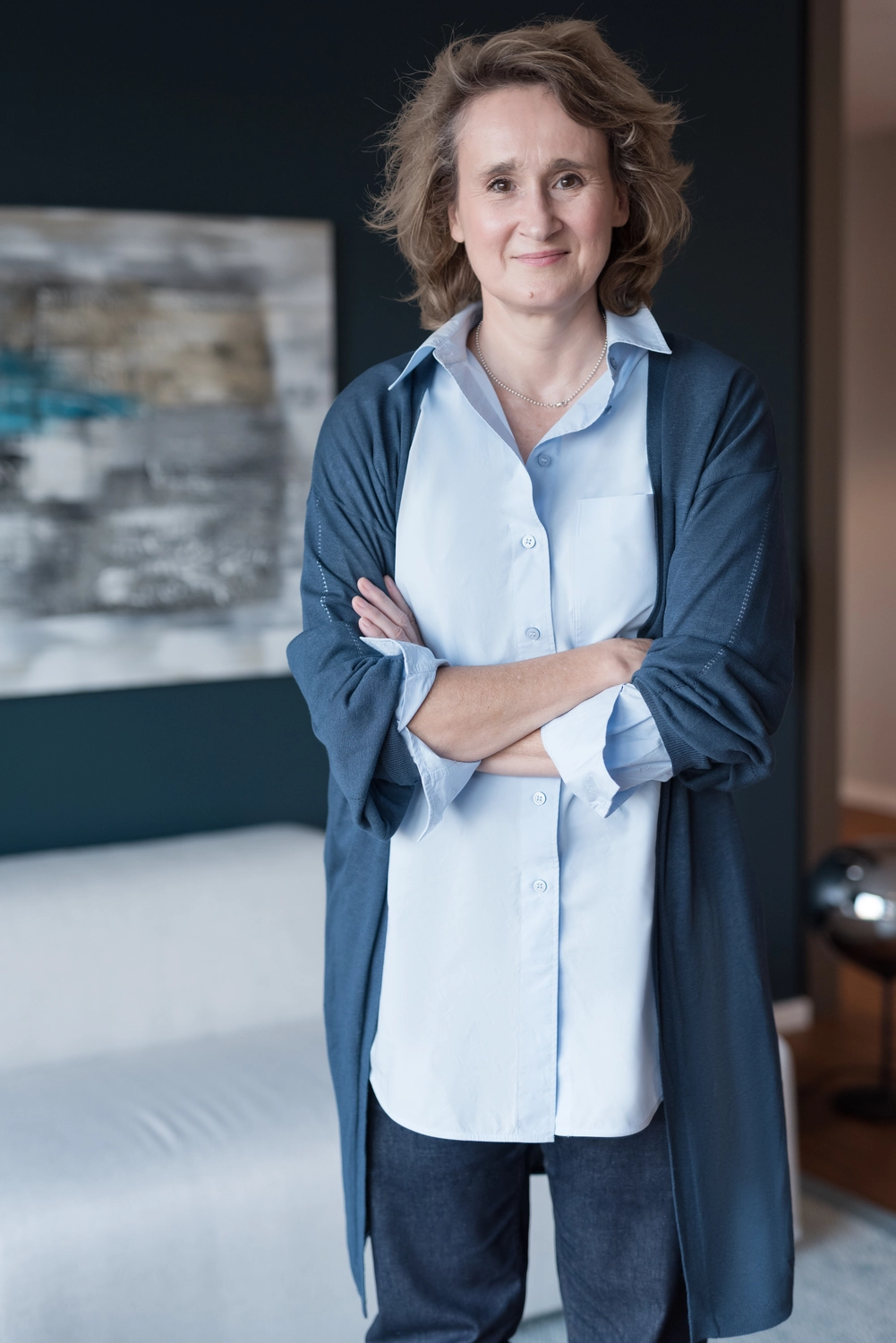 Portrait6: Christiane Schicker steht gerade und mit offenem Blick, und lächelnden Gesichtszügen vor dem Fotografen. Die Arme hat sie entspannt und selbstbewusst vor dem Bauch verschränkt. Sie ist in einem blauen Cardigan mit hellblauer Hembluse und einer blauen Hose gekleidet. Im Hintergrund sieht man ein helles modernes Sofa, darüber eine große Leinwand mit einem abstrakten Bild, das an einer dunkelblauen Wand hängt. Eine schicke Loungestehlampe ist neben dem Sofa auf dem hellgrauen Teppichboden noch zu erkennen. Das Ambiente strahlt elegantes, gleichzeitig wohnliches Businessflair eines Co-Working-Büros aus.