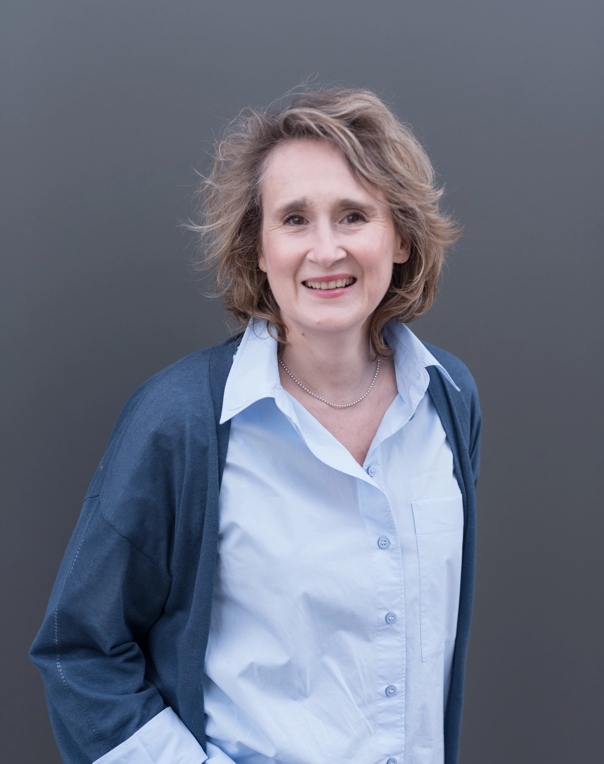 Portrait3 Christiane Schicker in blauem Cardigan und hellblauer Hemdbluse vor einem dunkelgrauen Hintergrund. Sie blickt mit offenem Lächeln in die Kamera, als ob sie sich mit dem Betrachter im Gespräch befinden würde.