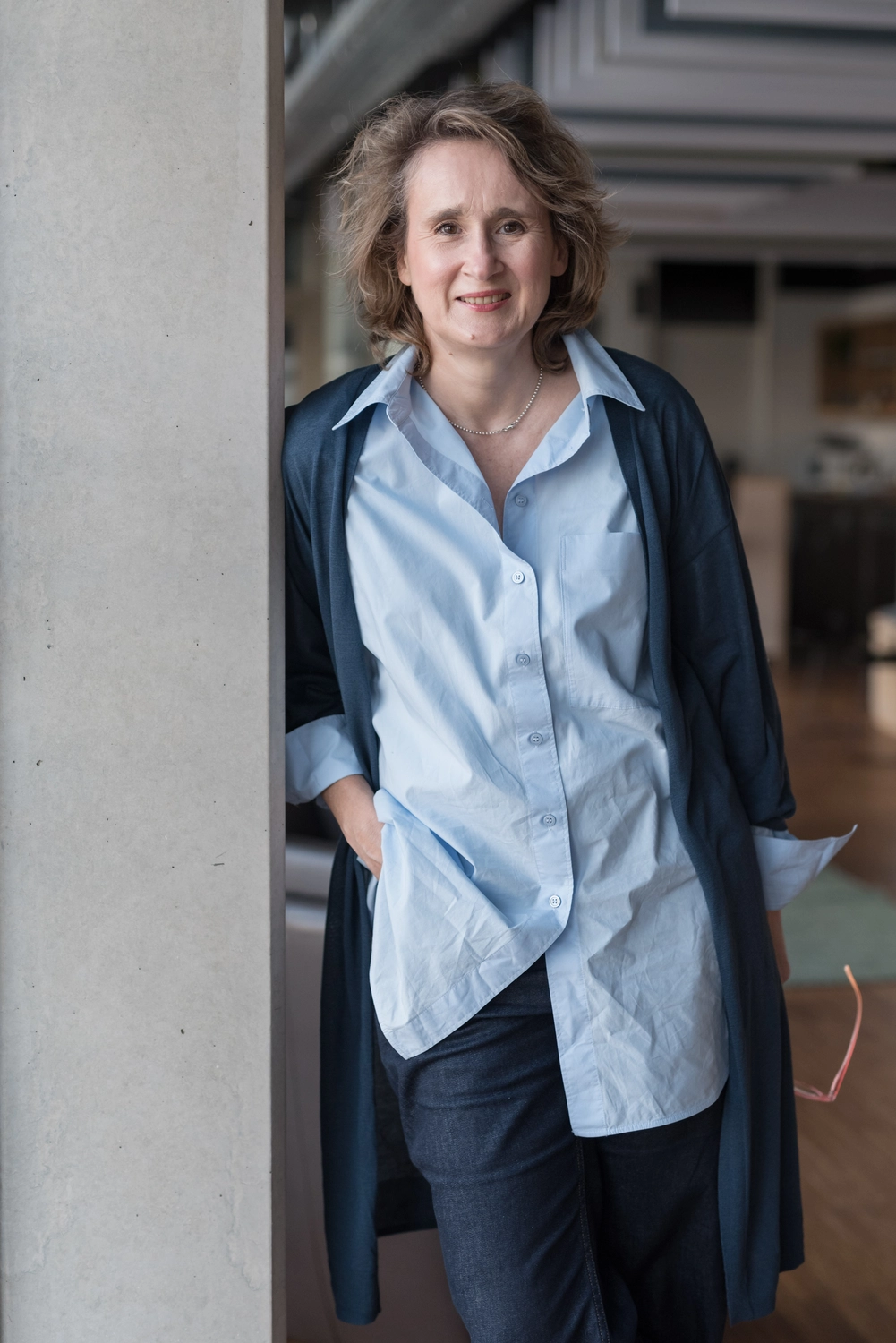 Portrait5: Christiane Schicker lehnt lässig an einem Betonpfeiler in einem loftähnlichen Raum mit viel Tiefe im Hintergrund. Sie lächelt entspannt in die Kamera. Der Arm, mit dem sie sich an den Pfeiler anlehnt, steckt in der Hosentasche. Sie ist gekleidet in einem blauen Cardigan mit hellblauer Hemdbluse.