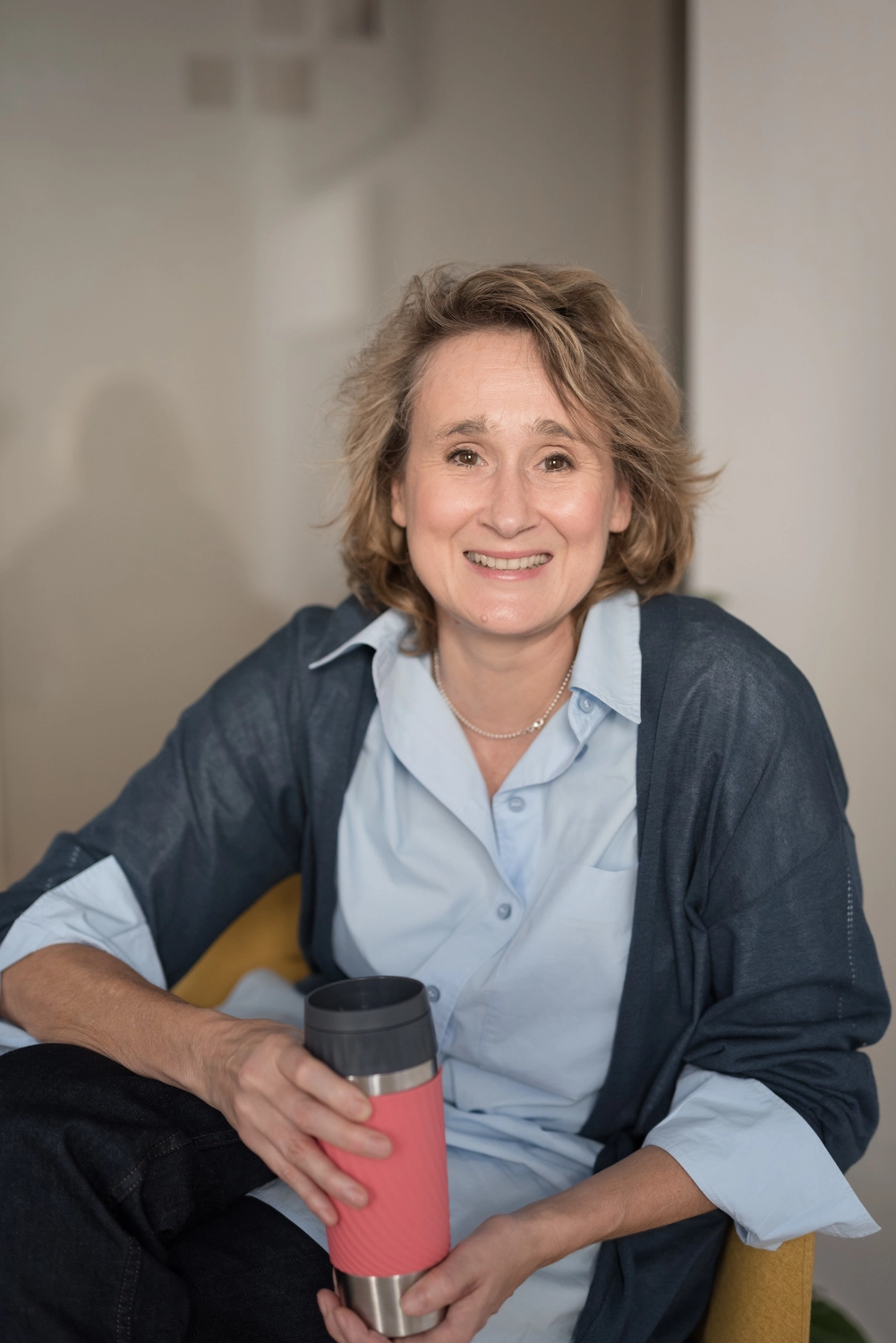 Portrait4: Christiane Schicker sitzt leicht nach vorne gebeugt gegenüber des Betrachters in einem Arbeitsraum. Sie lächelt offenherzig und hat die Beine übereinander geschlagen und die Arme lehnen locker auf den Oberschenkeln und der Sessellehne. Sie trägt einen einen blauen Cardigan über einer hellblauen Hemdbluse mit großen, umgekrempelten Manschetten auf halber Unterarmhöhe. Sie hält einen Thermotrinkbecher in den Händen, womit eine lockere, entspannte Gesprächsatmosphäre signalisiert wird.