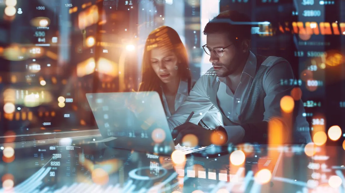Eine junge Frau und ein junger Mann mit Brille sitzen im Businesslook vor einem Laptop, in dem sie gemeinsam mit konzentrierten Gesichtern blicken. Sie sitzen im Dämmerlicht in einem Arbeitsraum vor dem Hintergrund einer Fensterkulisse mit großstädtischer Skyline. Das Bild wurde offensichtlich hinter einer Glaswand aufgenommen. Im Hintergrund blinken unscharf die Lichter aus den gegenüberliegenden Gebäudefenstern und Straßenlaternen im Fensterausschnitt. Im Vordergrund spiegeln sich weitere Lichter einer Raumbeleuchtung und blinkende Zahlen von anderen digitalen Bildschirmen in der Glaswand und überblenden die Szene mit den zwei Personen. Die großstädtische Skyline und die vielen elektrischen Impulse und blinkenden Bildschirme suggerieren futuristische und digitale Geschäftigkeit.