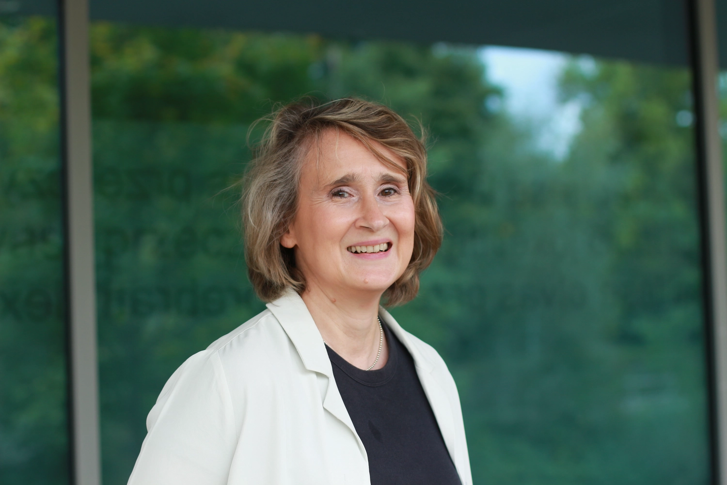 Portraitbild Christiane Schicker - Christiane blickt lächelnd mit leicht gedrehter Haltung in die Kamera. Sie trägt ein dunkles Rundhals-Tshirt unter einem weißen Jackett. Im Hintergrund sieht man sommerlich belaubte Hecken und einen Zipfel blauen Himmel.