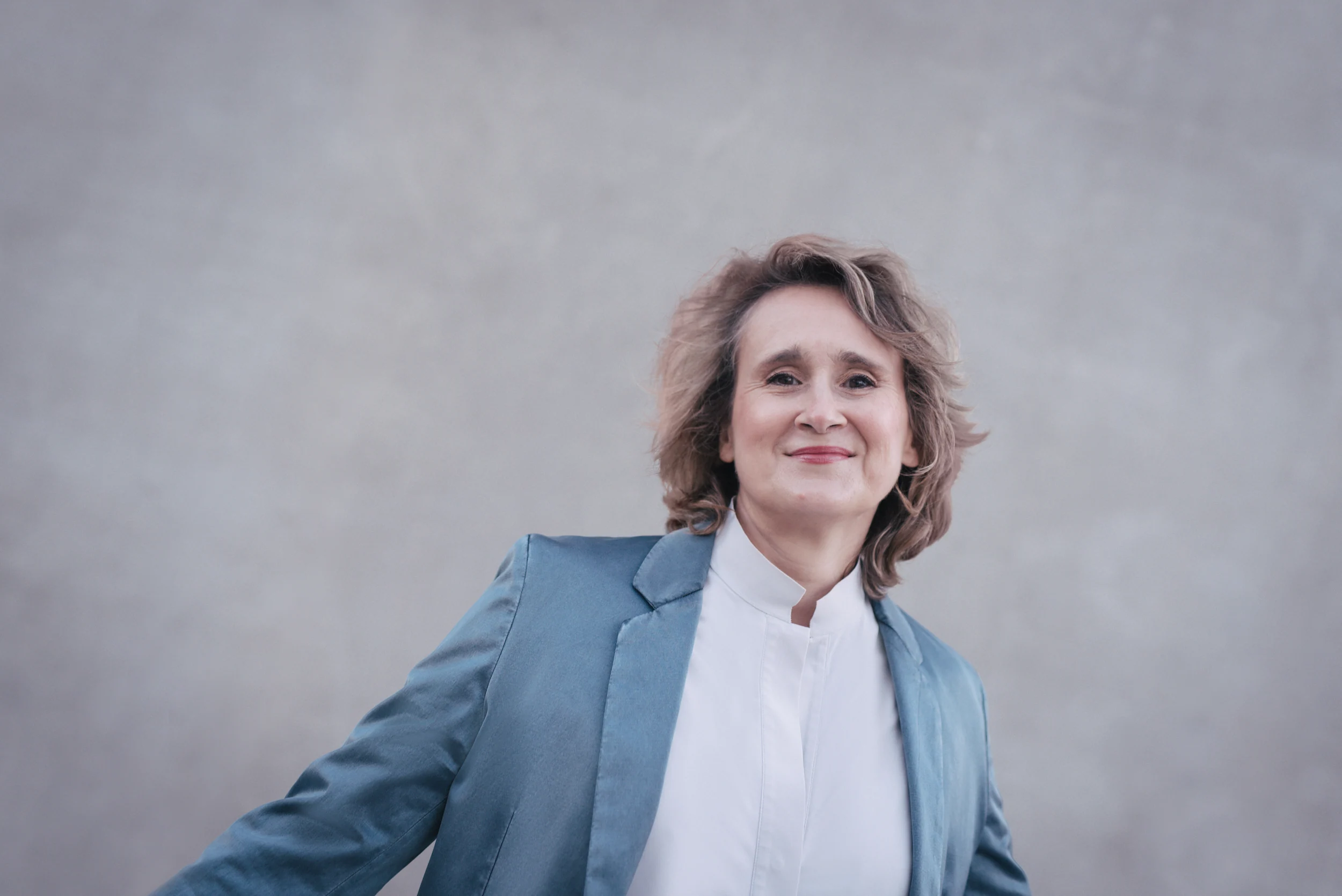 Portrait2 Christiane Schicker in blauem Blazer mit heller Hemdbluse vor grauem Hintergrund. Sie hat die Arme leicht ausgestreckt, als ob sie sich an einem Geländer festhält und blickt lächelnd von einer erhöhten Perspektive in die Kamera.